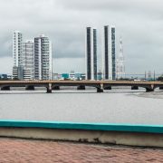 Vista do skyline de Recife, com edifícios, rio e ponte sob céu nublado.