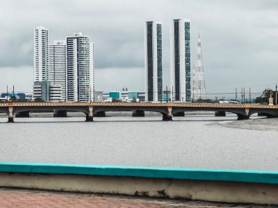 Vista do skyline de Recife, com edifícios, rio e ponte sob céu nublado.