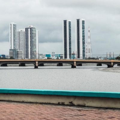 Vista do skyline de Recife, com edifícios, rio e ponte sob céu nublado.