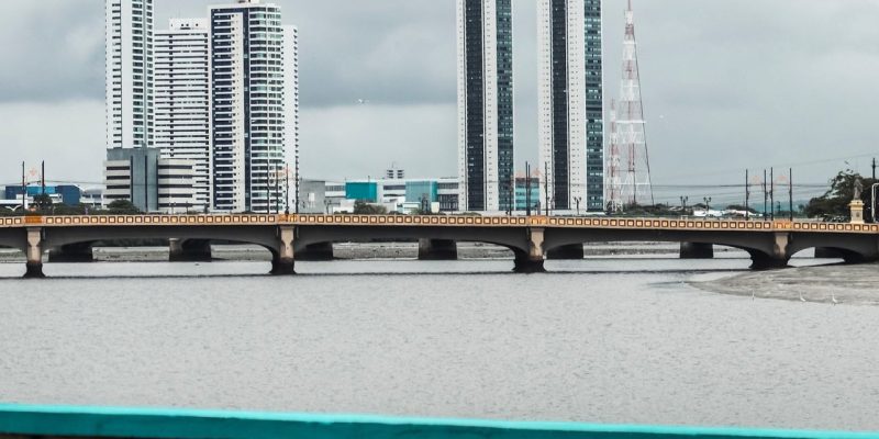 Vista do skyline de Recife, com edifícios, rio e ponte sob céu nublado.