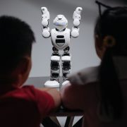 Duas crianças observam um robô humanoide sobre uma mesa em ambiente interno.