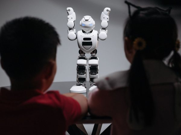 Duas crianças observam um robô humanoide sobre uma mesa em ambiente interno.