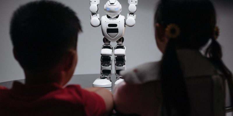 Duas crianças observam um robô humanoide sobre uma mesa em ambiente interno.